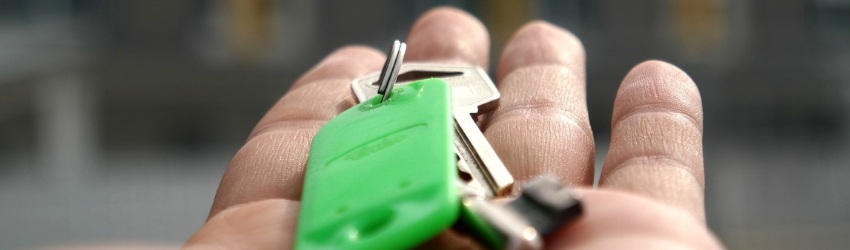 Elemento chiave della Vendita: mano di uomo tesa che regge un mazzo di chiavi con un portachiavi verde su sfondo di case sfocate