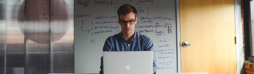 Regime Forfettario 2019: ragazzo che scrive al pc davanti ad un muro con schemi aziendali scritti sopra dentro un ufficio