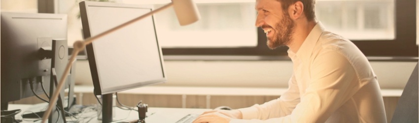 Uomo sorridente seduto ad una scrivania mentre lavora ad un computer dentro una stanza di ufficio. Sullo sfondo una finestra 