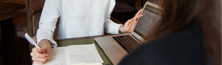 Vendi di più imparando ad auto-valutarti: venditrice seduta a un tavol con dei documenti davanti che conversando con una cliente le mostra qualcosa sullo schermo di un computer